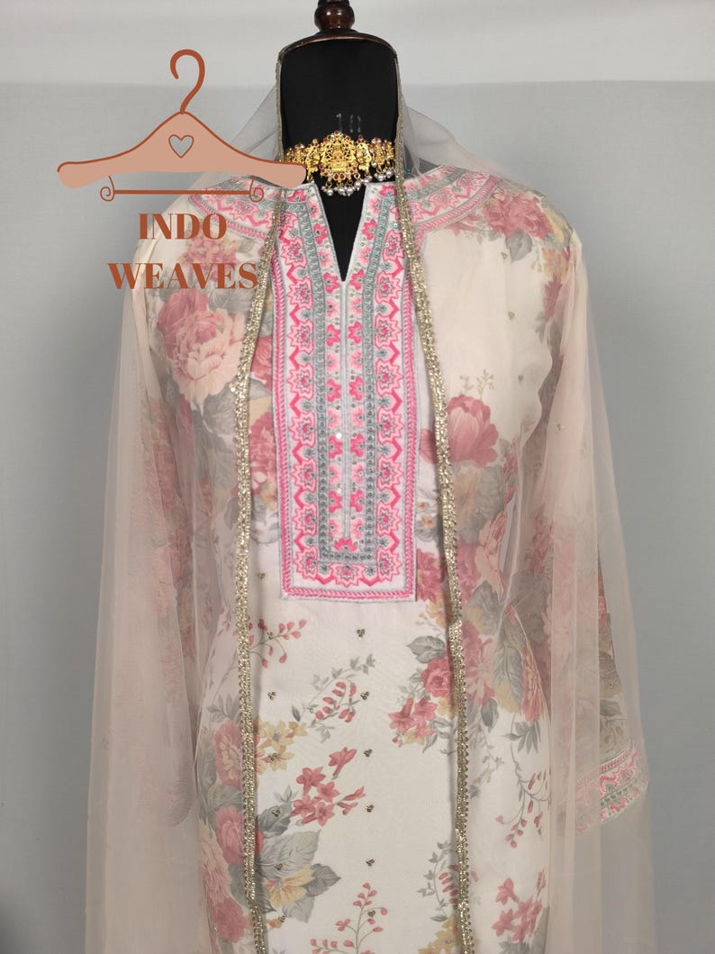 K&ouml;nnte beinhalten: Ein Salwar Kameez mit floralem Muster und passendem Dupatta. Der cremefarbene Stoff weist ein gro&szlig;es Muster aus rosa und roten Rosen auf. Der Ausschnitt und die Kanten sind mit Stickereien und goldenen Zierleisten verziert. Das Outfit wird auf einer Schaufenquin mit einer goldenen Halskette pr&auml;sentiert.