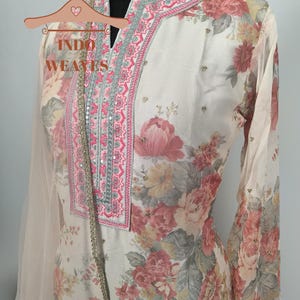 K&ouml;nnte beinhalten: Cremefarbenes Outfit mit floralem Druck und rosa und grauen Blumenmustern. Das Outfit hat einen V-Ausschnitt mit rosa und silberner Stickerei. Die &Auml;rmel sind lang und das Outfit ist mit kleinen, funkelnden Verzierungen versehen. Der Text "INDO WEAVES" ist sichtbar.