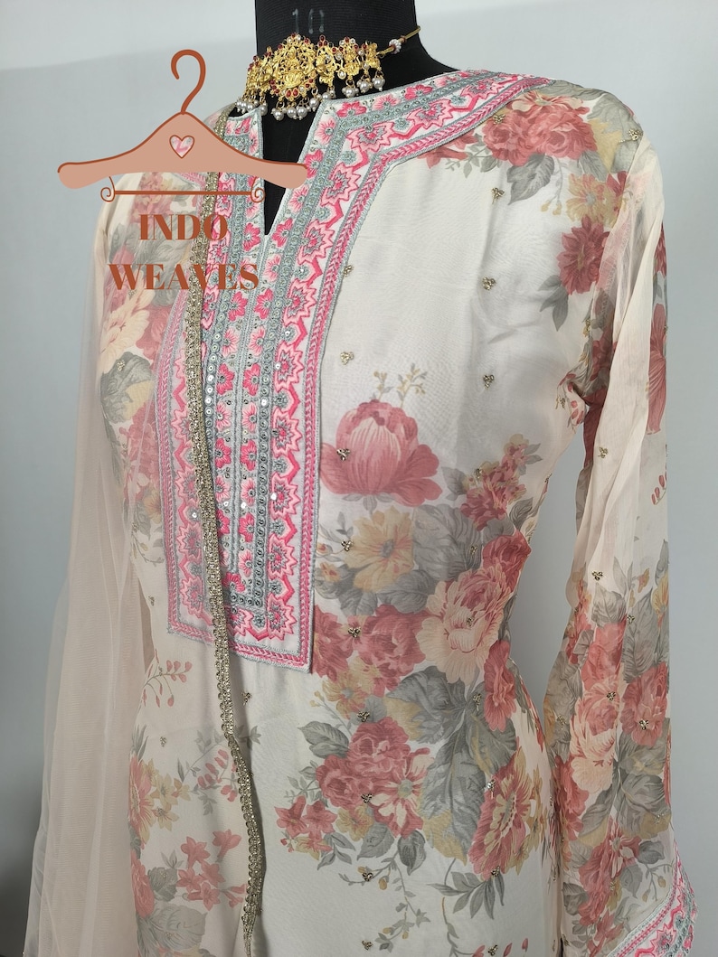 K&ouml;nnte beinhalten: Cremefarbenes Outfit mit floralem Druck und rosa und grauen Blumenmustern. Das Outfit hat einen V-Ausschnitt mit rosa und silberner Stickerei. Die &Auml;rmel sind lang und das Outfit ist mit kleinen, funkelnden Verzierungen versehen. Der Text "INDO WEAVES" ist sichtbar.