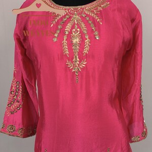 Pu&ograve; includere: Una tunica rosa acceso con ricami dorati intricati. La tunica presenta maniche lunghe con polsini dettagliati e abbellimenti ornati intorno allo scollo e al corpetto. L'indumento &egrave; esposto su un manichino, mettendo in risalto il suo design elegante.