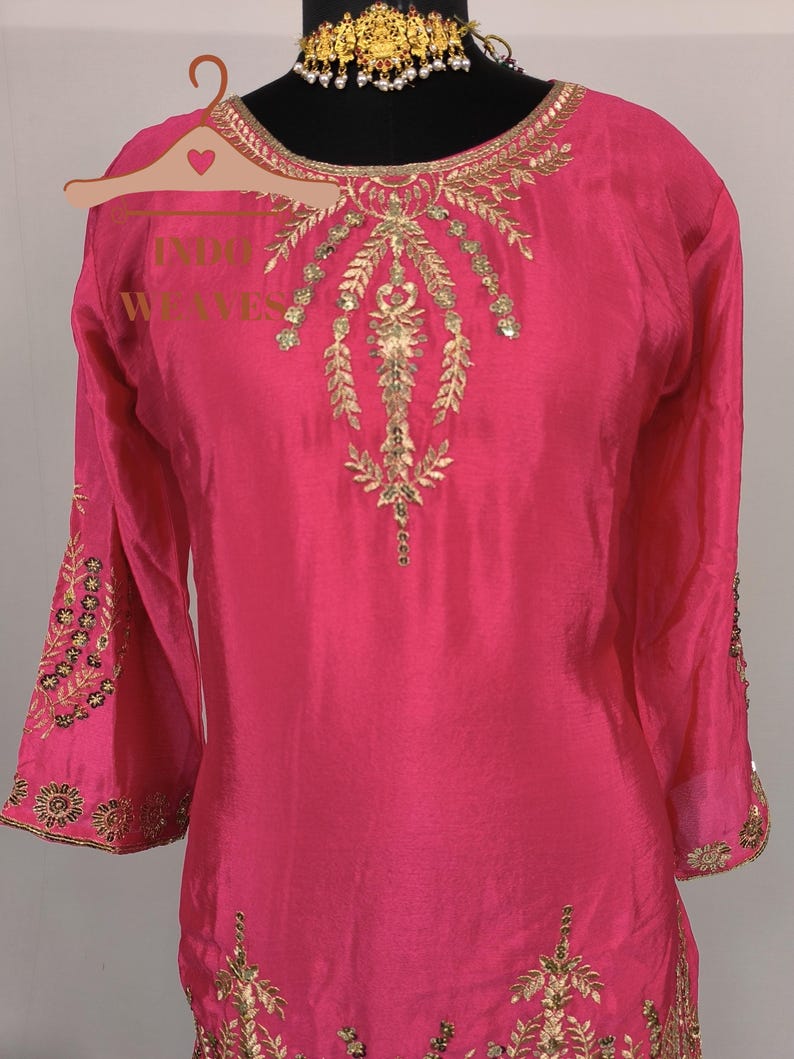 Pu&ograve; includere: Una tunica rosa acceso con ricami dorati intricati. La tunica presenta maniche lunghe con polsini dettagliati e abbellimenti ornati intorno allo scollo e al corpetto. L'indumento &egrave; esposto su un manichino, mettendo in risalto il suo design elegante.