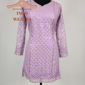 Puede incluir: Un vestido t&uacute;nica lavanda con mangas largas y cuello en V, adornado con lentejuelas doradas en un patr&oacute;n geom&eacute;trico. El vestido presenta bordes festoneados y se exhibe en un maniqu&iacute;. Tambi&eacute;n se ve un collar gargantilla dorado.
