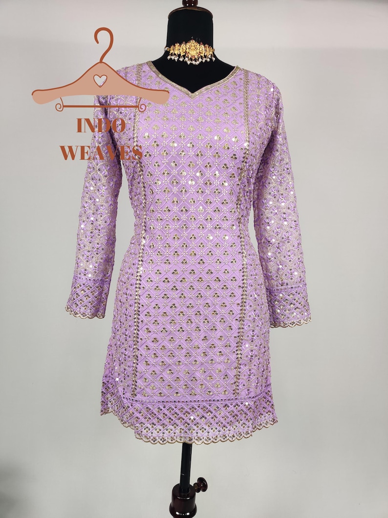 Puede incluir: Un vestido t&uacute;nica lavanda con mangas largas y cuello en V, adornado con lentejuelas doradas en un patr&oacute;n geom&eacute;trico. El vestido presenta bordes festoneados y se exhibe en un maniqu&iacute;. Tambi&eacute;n se ve un collar gargantilla dorado.