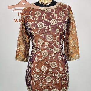 May include: A brown and purple tunic with intricate floral embroidery. The three-quarter sleeves and neckline are trimmed with gold-colored accents. A gold-colored necklace is displayed on the mannequin. The tunic is displayed on a black mannequin stand.