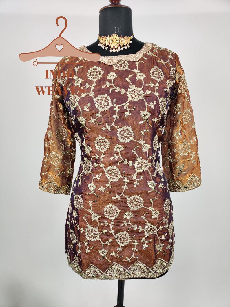 May include: A brown and purple tunic with intricate floral embroidery. The three-quarter sleeves and neckline are trimmed with gold-colored accents. A gold-colored necklace is displayed on the mannequin. The tunic is displayed on a black mannequin stand.