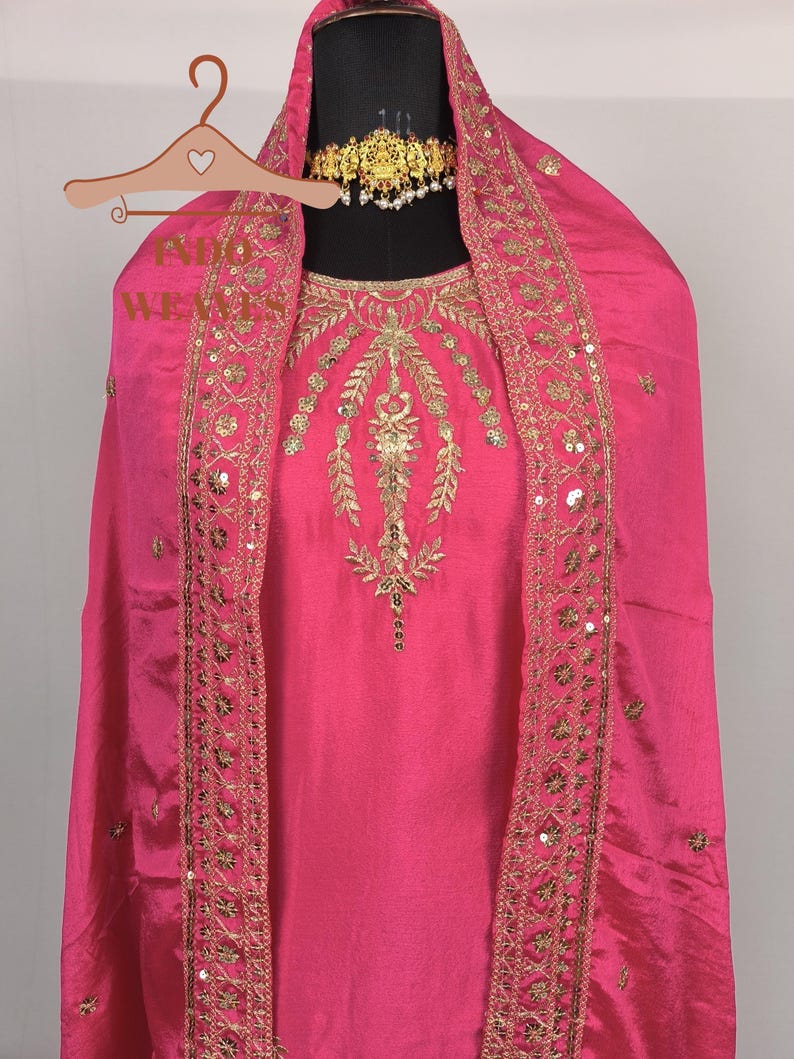 Pu&ograve; includere: Un vivace completo salwar kameez rosa. L'outfit presenta intricati ricami dorati e abbellimenti di paillettes lungo lo scollo e i bordi. Un dupatta abbinato &egrave; drappeggiato sulle spalle. Una collana girocollo dorata aggiunge un tocco di eleganza.