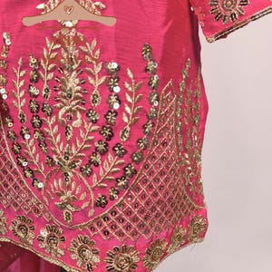 Pu&ograve; includere: Primo piano di un vivace capo rosa con ricami dorati intricati. Il design presenta motivi floreali e a foglie, impreziositi da paillettes. I bordi del capo sono rifiniti con dettagli dorati, creando un aspetto lussuoso.