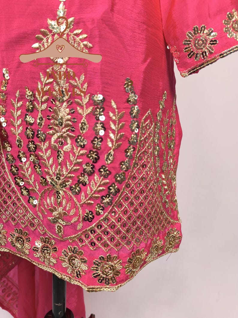 Pu&ograve; includere: Primo piano di un vivace capo rosa con ricami dorati intricati. Il design presenta motivi floreali e a foglie, impreziositi da paillettes. I bordi del capo sono rifiniti con dettagli dorati, creando un aspetto lussuoso.