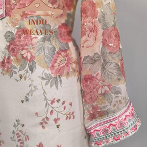 K&ouml;nnte beinhalten: Ein wei&szlig;es Kleidungsst&uuml;ck mit floralem Druck mit rosa, roten und grauen Blumen. Der lange &Auml;rmel hat eine dekorative rosa und gr&uuml;ne Bord&uuml;re. Das Kleidungsst&uuml;ck ist mit kleinen goldenen Pailletten verziert. Der Text "INDO WEAVES" ist sichtbar.