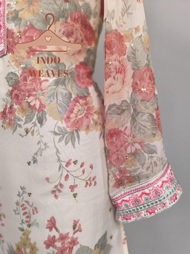 K&ouml;nnte beinhalten: Ein wei&szlig;es Kleidungsst&uuml;ck mit floralem Druck mit rosa, roten und grauen Blumen. Der lange &Auml;rmel hat eine dekorative rosa und gr&uuml;ne Bord&uuml;re. Das Kleidungsst&uuml;ck ist mit kleinen goldenen Pailletten verziert. Der Text "INDO WEAVES" ist sichtbar.