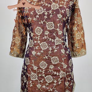 May include: A brown and purple tunic with floral embroidery. The tunic has three-quarter sleeves and a scalloped hem. The floral embroidery is in a cream color and covers the front and sleeves of the tunic.