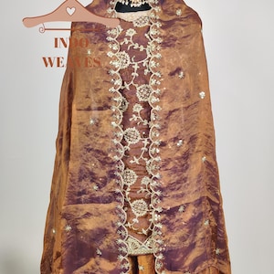 May include: A formal ensemble in shades of bronze and deep purple, featuring intricate embroidery and embellishments. The outfit includes a draped shawl, a skirt, and a matching top, all displayed on a mannequin. A gold-toned necklace adds a touch of elegance.