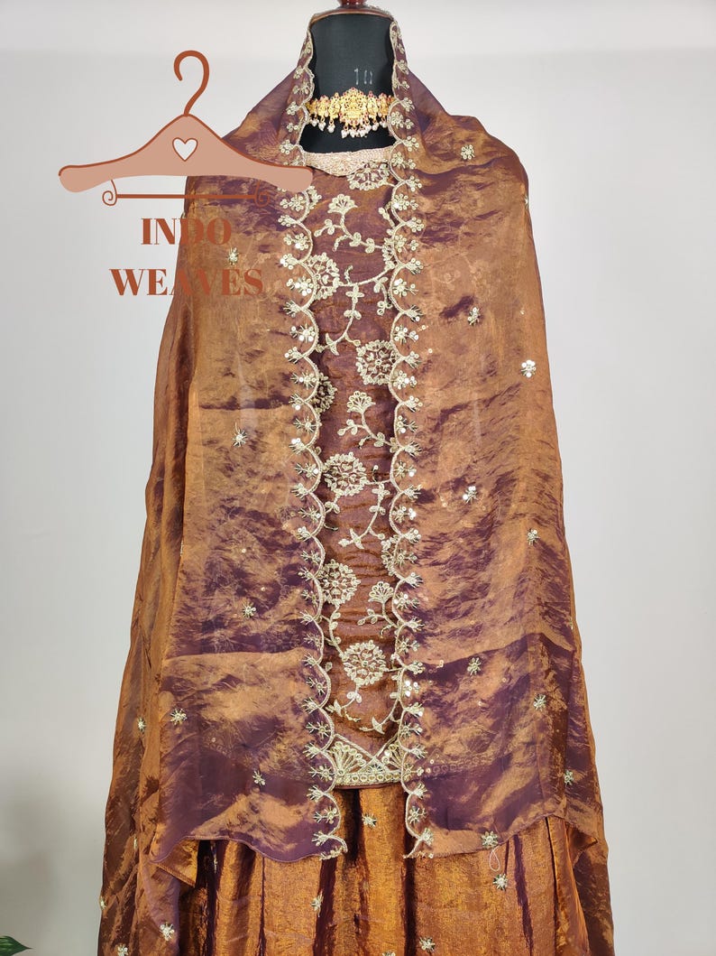 May include: A formal ensemble in shades of bronze and deep purple, featuring intricate embroidery and embellishments. The outfit includes a draped shawl, a skirt, and a matching top, all displayed on a mannequin. A gold-toned necklace adds a touch of elegance.
