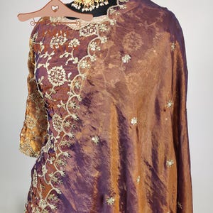 May include: A two-piece ensemble in shades of bronze and purple, featuring intricate gold embroidery and embellishments. The outfit includes a long-sleeved top and a draped shawl. A gold-toned necklace with red and white accents is also visible.