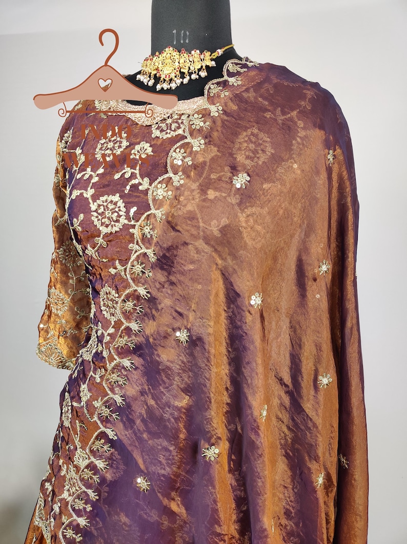 May include: A two-piece ensemble in shades of bronze and purple, featuring intricate gold embroidery and embellishments. The outfit includes a long-sleeved top and a draped shawl. A gold-toned necklace with red and white accents is also visible.