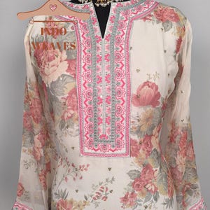K&ouml;nnte beinhalten: Cremefarbene Tunika mit floralem Print und langen &Auml;rmeln. Die Tunika hat einen V-Ausschnitt mit rosa und silberner Stickerei. Das Blumenmuster umfasst rosa, rote und gelbe Blumen mit gr&uuml;nen Bl&auml;ttern. Der Text "INDO WAVES" ist auf einem Kleiderb&uuml;gel zu sehen.