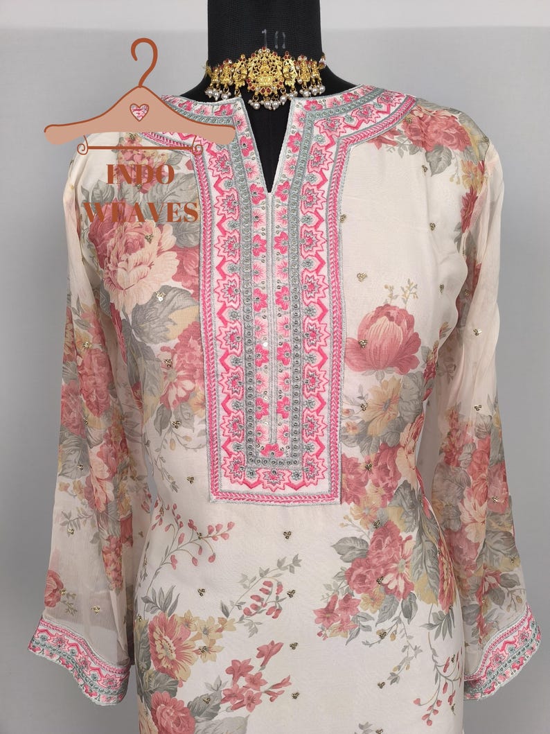 K&ouml;nnte beinhalten: Cremefarbene Tunika mit floralem Print und langen &Auml;rmeln. Die Tunika hat einen V-Ausschnitt mit rosa und silberner Stickerei. Das Blumenmuster umfasst rosa, rote und gelbe Blumen mit gr&uuml;nen Bl&auml;ttern. Der Text "INDO WAVES" ist auf einem Kleiderb&uuml;gel zu sehen.