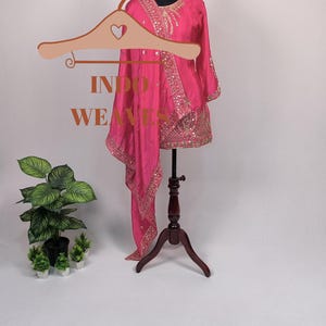 Pu&ograve; includere: Un vivace completo indiano rosa con ricami dorati e paillettes. L'outfit include una tunica e una sciarpa abbinata, esposto su un manichino. Il testo "INDO WEAVES" &egrave; visibile su un appendiabiti.