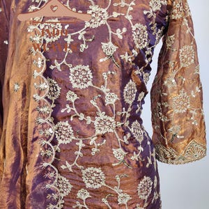 May include: A plum-colored, long-sleeved garment with intricate gold floral embroidery. The fabric has a shimmering, iridescent quality. The sleeves are flared at the wrist, and the neckline is trimmed with gold embellishments. The embroidery is detailed and covers the garment.