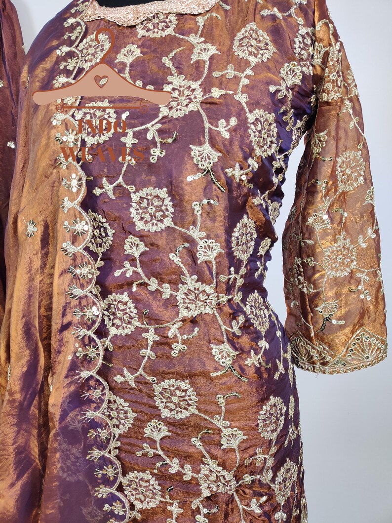 May include: A plum-colored, long-sleeved garment with intricate gold floral embroidery. The fabric has a shimmering, iridescent quality. The sleeves are flared at the wrist, and the neckline is trimmed with gold embellishments. The embroidery is detailed and covers the garment.