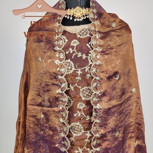 May include: A brown and purple silk garment with intricate floral embroidery and sequin accents. The fabric drapes over a mannequin, showcasing a gold-toned necklace. The garment features a detailed, embellished neckline and a flowing design.