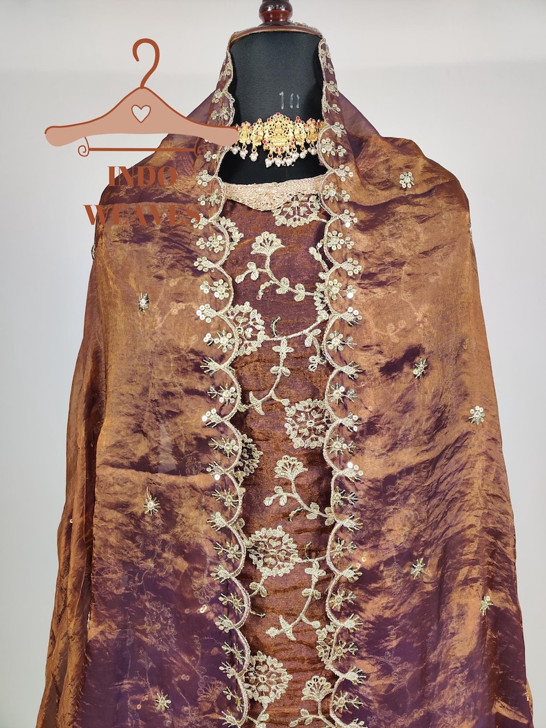 May include: A brown and purple silk garment with intricate floral embroidery and sequin accents. The fabric drapes over a mannequin, showcasing a gold-toned necklace. The garment features a detailed, embellished neckline and a flowing design.