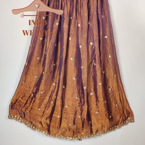 May include: A floor-length skirt in shades of bronze and purple, hanging on a wooden hanger. The skirt is made of a shimmering fabric with a subtle sheen and is adorned with small, embroidered floral accents. The hemline is finished with a delicate, beaded trim.