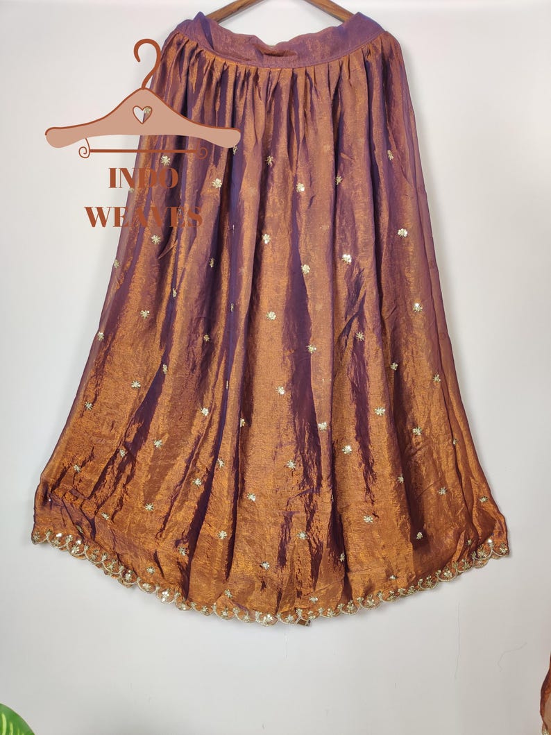 May include: A floor-length skirt in shades of bronze and purple, hanging on a wooden hanger. The skirt is made of a shimmering fabric with a subtle sheen and is adorned with small, embroidered floral accents. The hemline is finished with a delicate, beaded trim.