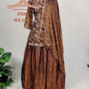 May include: A brown Indian ethnic wear ensemble, including a long-sleeved top and a flowing skirt, displayed on a mannequin. The outfit features intricate embroidery and embellishments. A matching shawl drapes over the shoulder. The text "INDO WEAR" is visible.