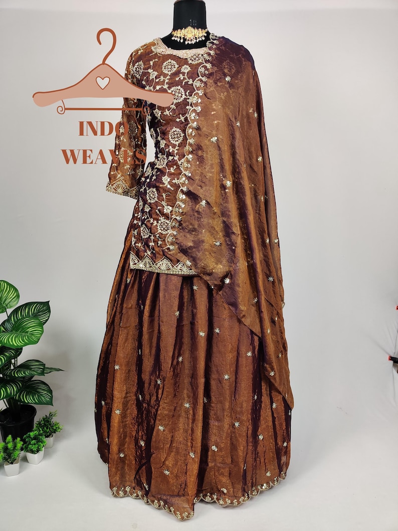 May include: A brown Indian ethnic wear ensemble, including a long-sleeved top and a flowing skirt, displayed on a mannequin. The outfit features intricate embroidery and embellishments. A matching shawl drapes over the shoulder. The text "INDO WEAR" is visible.
