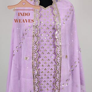 Puede incluir: Conjunto salwar kameez lila claro con detalles de lentejuelas y bordados dorados. El atuendo incluye una t&uacute;nica, una bufanda a juego y un collar gargantilla dorado. El texto "INDO WEAVES" es visible en una percha.