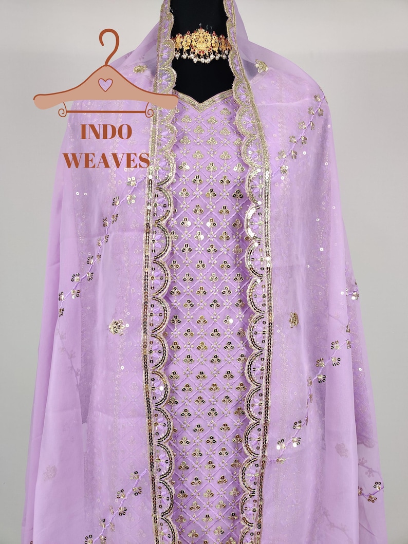 Puede incluir: Conjunto salwar kameez lila claro con detalles de lentejuelas y bordados dorados. El atuendo incluye una t&uacute;nica, una bufanda a juego y un collar gargantilla dorado. El texto "INDO WEAVES" es visible en una percha.