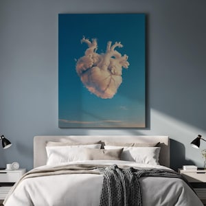 May include: A canvas print featuring a surreal image of a heart-shaped cloud floating in a blue sky. The artwork is displayed above a bed with white bedding and a gray throw blanket. The room has a modern aesthetic.