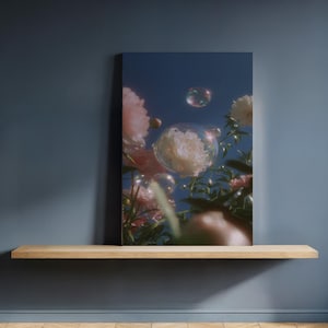 Puede incluir: Impresión en lienzo que muestra flores rosas y blancas con burbujas flotando contra un cielo azul. La obra de arte se exhibe en una estantería de madera contra una pared azul oscuro. La imagen evoca fantasía.