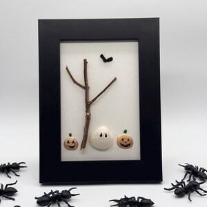 Puede incluir: Una obra de arte enmarcada con temática de Halloween que presenta una rama de árbol, un fantasma de concha y dos decoraciones de calabaza sonrientes. Hormigas de plástico negro rodean el marco. La obra está sobre un fondo blanco, con un marco negro.