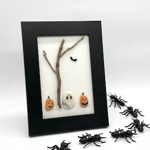 Puede incluir: Una obra de arte enmarcada con temática de Halloween que presenta un árbol hecho de ramitas, un fantasma y dos calabazas talladas. Hormigas de plástico negro dispuestas en fila, que conducen al marco. El marco es negro.