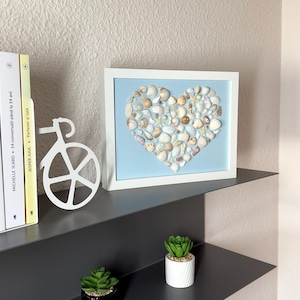 Puede incluir: Obra de arte enmarcada con forma de corazón hecha de conchas marinas sobre un fondo azul claro. El marco blanco está sobre una estantería gris oscuro, junto con un adorno blanco en forma de bicicleta y pequeñas suculentas en macetas.
