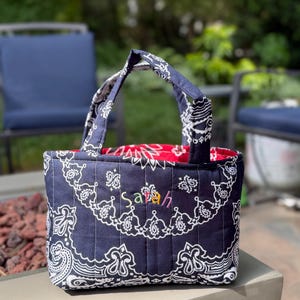 May include: A quilted fabric tote bag featuring a navy blue and white paisley bandana pattern. The bag has two matching handles and the name Sarah embroidered in multi-coloured thread on the front. The interior lining is a solid bright red fabric.