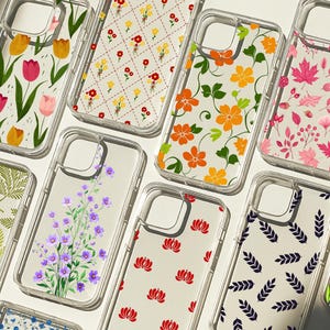 May include: A selection of clear phone cases showcasing floral designs. The cases display patterns of tulips, orange flowers, red flowers, and purple flowers. Other cases feature leaf and geometric patterns. The cases are made of clear plastic.