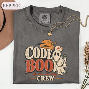May include: A charcoal gray t-shirt featuring a graphic with the text CODE BOO CREW. The design includes a ghost wearing a nurse cap and holding a stethoscope, an orange moon, and a red heartbeat line. The shirt has a Comfort Colors brand label.