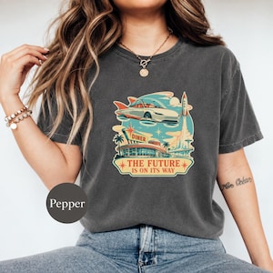 May include: A charcoal gray t-shirt featuring a graphic print of a retro-futuristic scene with a flying car, a rocket, and a diner. The text reads "DINER" and "THE FUTURE IS ON ITS WAY" in orange and yellow lettering with star accents.