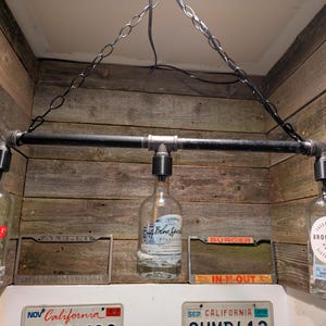 May include: A unique, industrial-style light fixture made from black pipes and repurposed liquor bottles. The fixture is suspended from a chain and features three light bulbs. The bottles have labels from various brands. The background is a wooden wall.