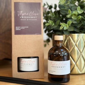 May include: A brown glass reed diffuser bottle with a wooden cap and a white label that reads "Thyme Olive + Bergamot". A matching box and a small container are also visible, along with black diffuser sticks. The scent is "Burnt Blossom".