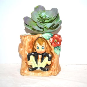 May include: A ceramic planter shaped like a tree stump with a figure sitting inside. The figure has light skin, brown hair, and wears a black jacket with yellow pants. A red flower with green leaves is attached to the side, and a green succulent sits on top.