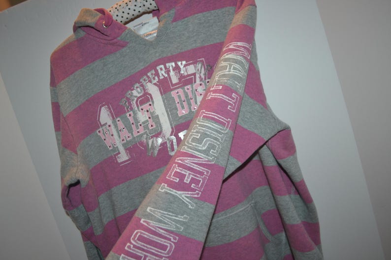 Pu&ograve; includere: Una felpa con cappuccio a righe grigie e rosa con la scritta Walt Disney World stampata in argento sulla manica e sul davanti. La grafica frontale include lettere argentate e piccole borchie incastonate. Il cappuccio ha una fodera a pois bianchi e neri.