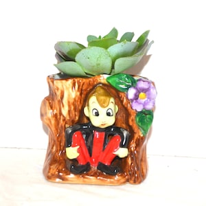 Puede incluir: Una maceta de cerámica con forma de tronco de árbol marrón que presenta una pequeña figura con cara bronceada, camisa negra y pantalones rojos sentada dentro. Una flor morada con hojas verdes está unida al lado y una suculenta verde en la parte superior.