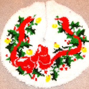May include: A white, round Christmas tree skirt with a festive design. The skirt features a red ribbon, green garland, yellow accents, and a red bow. The design is set against a white background.