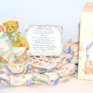 May include: A collectible "Cherished Teddies" figurine featuring a teddy bear and a doll in a wagon. The figurine is on a crocheted doily. A certificate of adoption and the original box are also visible.