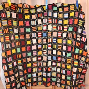 Könnte beinhalten: Eine handgehäkelte Decke mit einem Gitter aus kleinen, bunten Quadraten in Gelb-, Orange-, Rot-, Blau-, Grün- und Rosatönen, die alle von einem durchgehenden schwarzen Garnnetz umrandet sind, das jedes einzelne Quadrat miteinander verbindet.