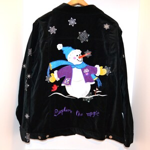 May include: Black corduroy jacket with a snowman applique and snowflake embellishments. The snowman wears a blue hat and scarf, a purple jacket, and yellow gloves. The phrase "Capture the magic" is embroidered in purple.